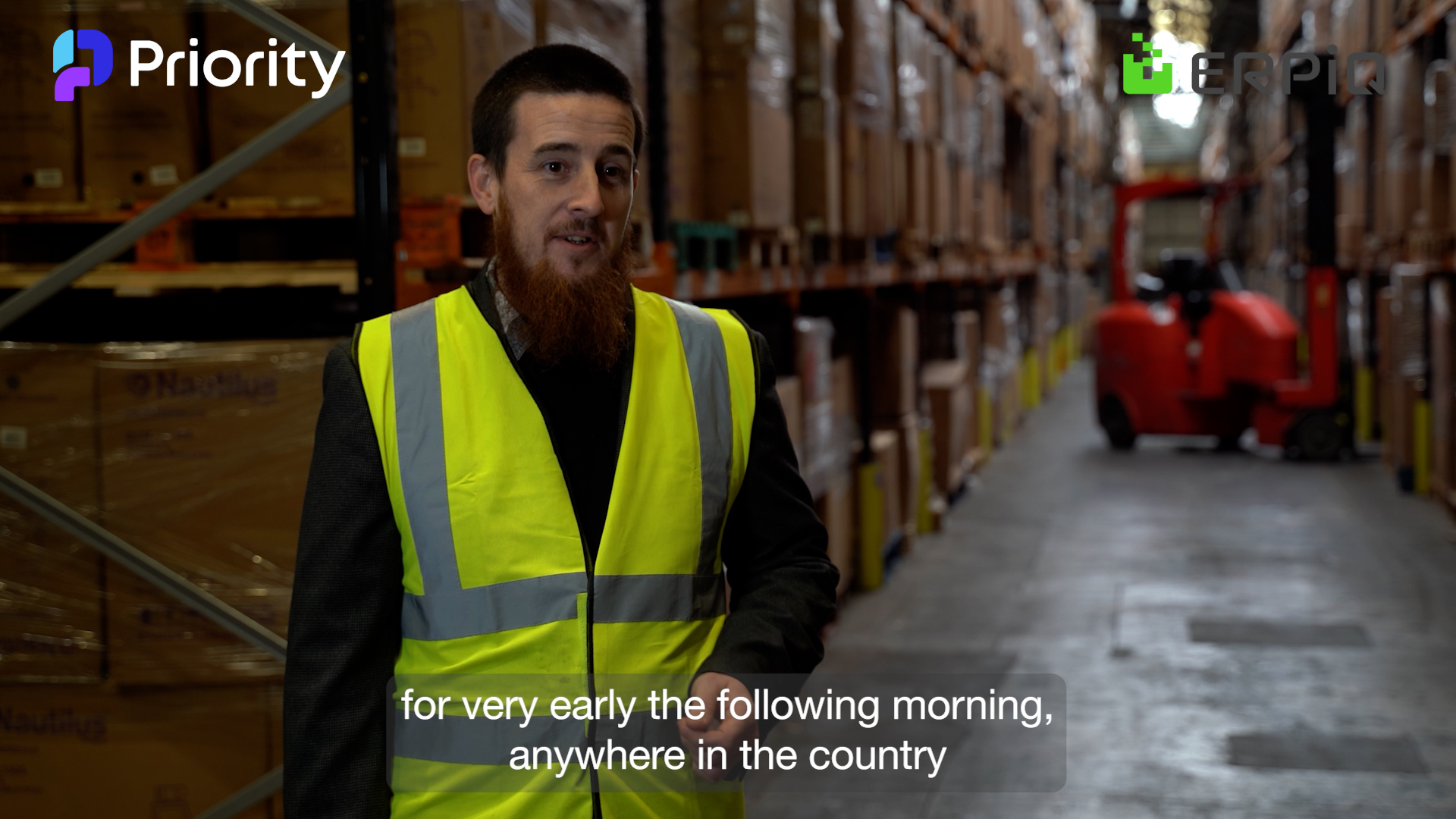 A man with a beard wearing a high-visibility vest stands in a warehouse aisle with shelves of products, and a red forklift visible in the background.